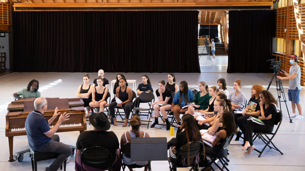 A group of performers sit in chairs grouped around a piano in a studio.