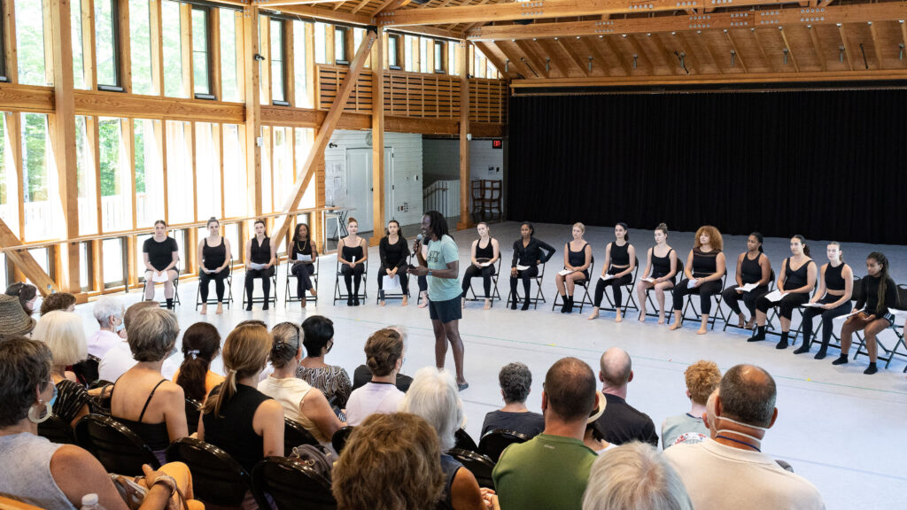 Jeffrey Page speaks to an audience. Behind him, there is a semi-circle of performers sitting in chairs. They are all wearing black.