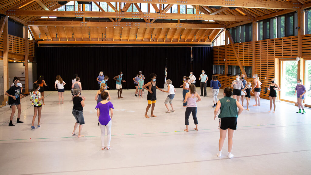 A group of dancers in a studio face inward toward Leonardo Sandoval in a loose circle.