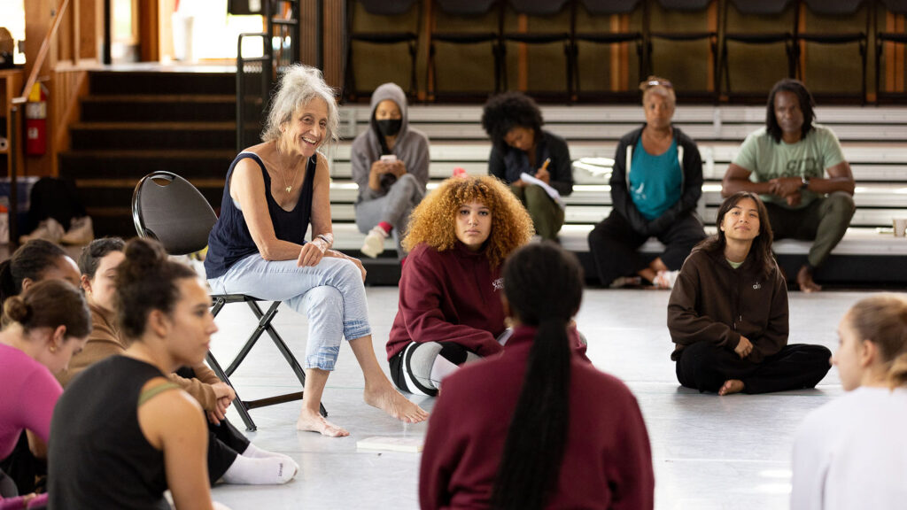 Liz Lerman sits on a chair surrounded by performers sitting on the ground.