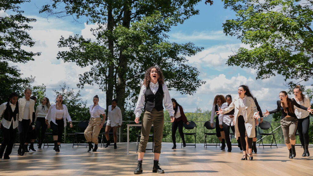 A performer stands and sings at the front of an outdoor stage, with a group of performers loosely circled behind. They wear colonial-inspired garb.