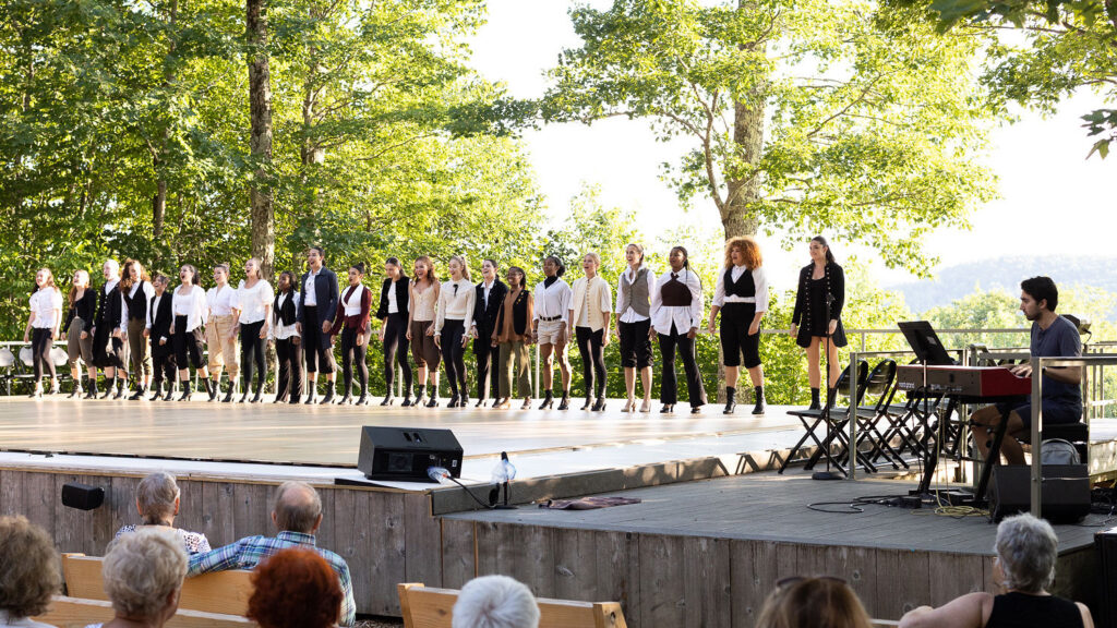 A line of performers dressed in colonial-inspired garb face an audience on an outdoor stage. A pianist sits off to the side.