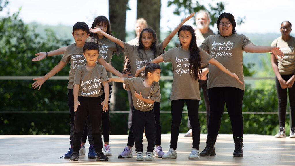 A group of young dancers on an outdoor stage wearing matching beige shirts that say "Dance for Social Justice."