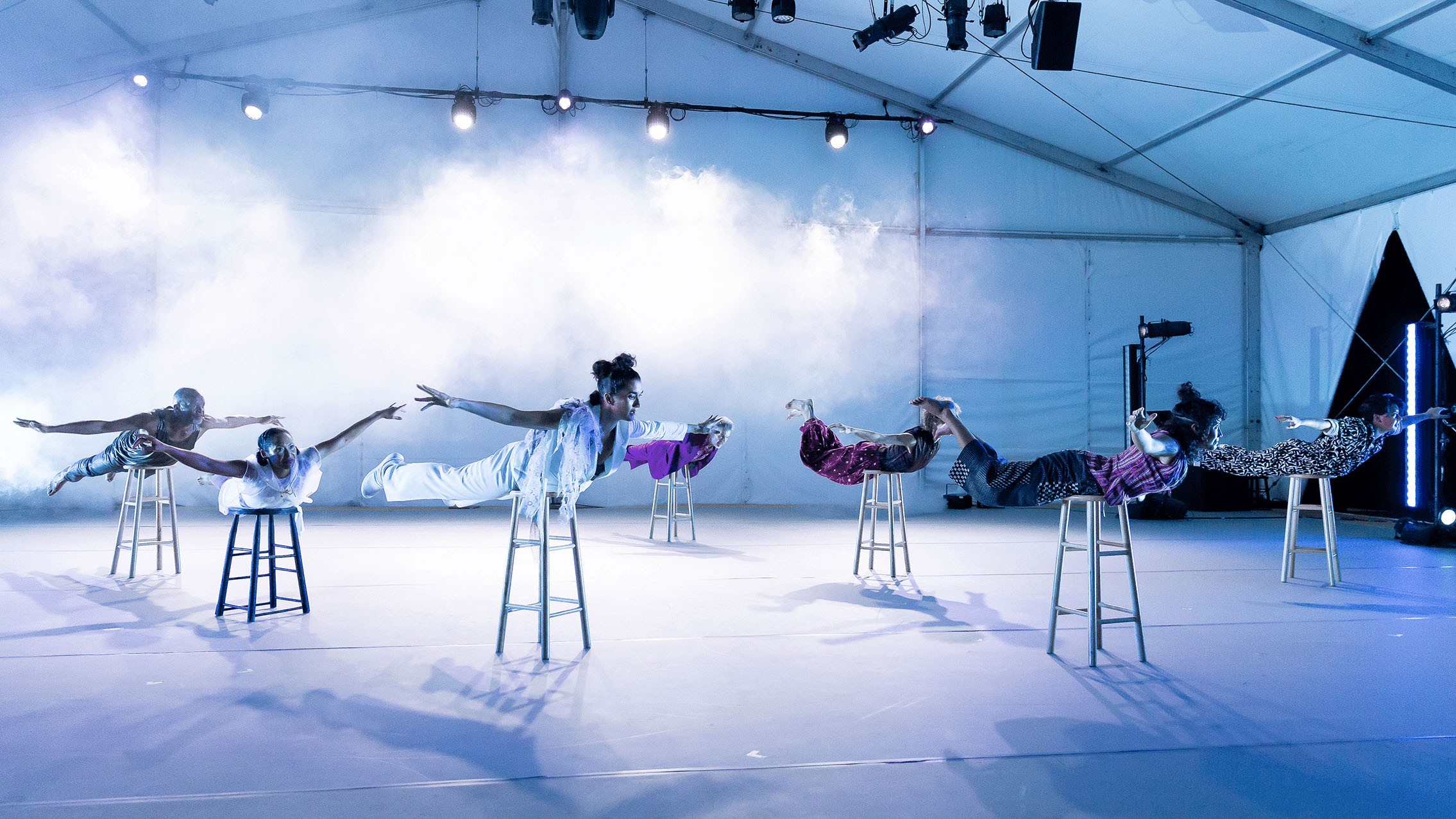 The cast of Liz Lerman laying on a stool with their bodies stretched out resembling flight.