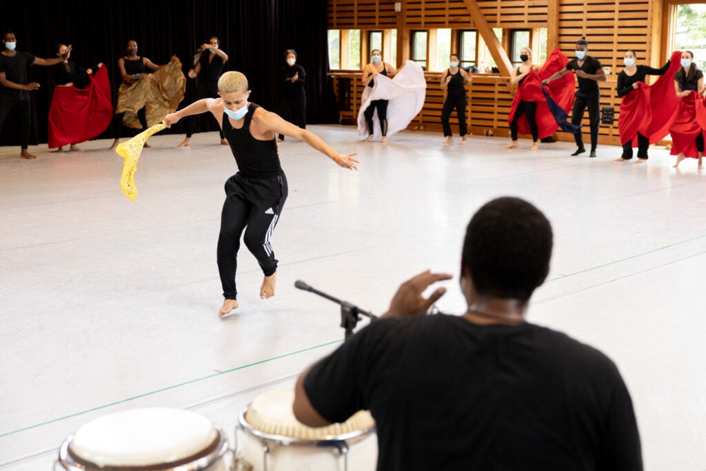 A dancer faces a drummer in a studio. The dancer spreads his arms, holding a yellow cloth in his right hand. He is in motion and looks as if he is about to whip the cloth.