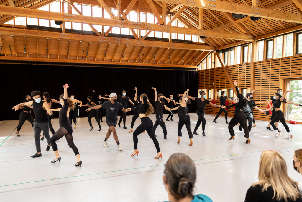 A group of dancers performing the Latin Hustle, where they partner dance, for an audience in a studio. The dancers are wearing all black and paired in groups of two.