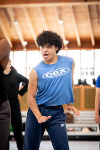 A dancer in a blue t-shirt opens his mouth and shifts his shoulders facing the camera. His arms are slightly bent with wrists flexed.