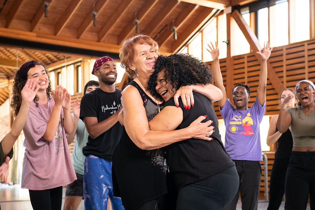 Maria Torres hugs Cleo Parker Robinson. A group of dancers stand around them and clap.
