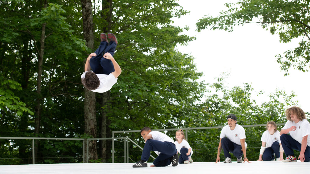 A group of dancers on an outdoor stage. One dancer in the front is mid backflip, while the dancers in the back crouch low and look on.