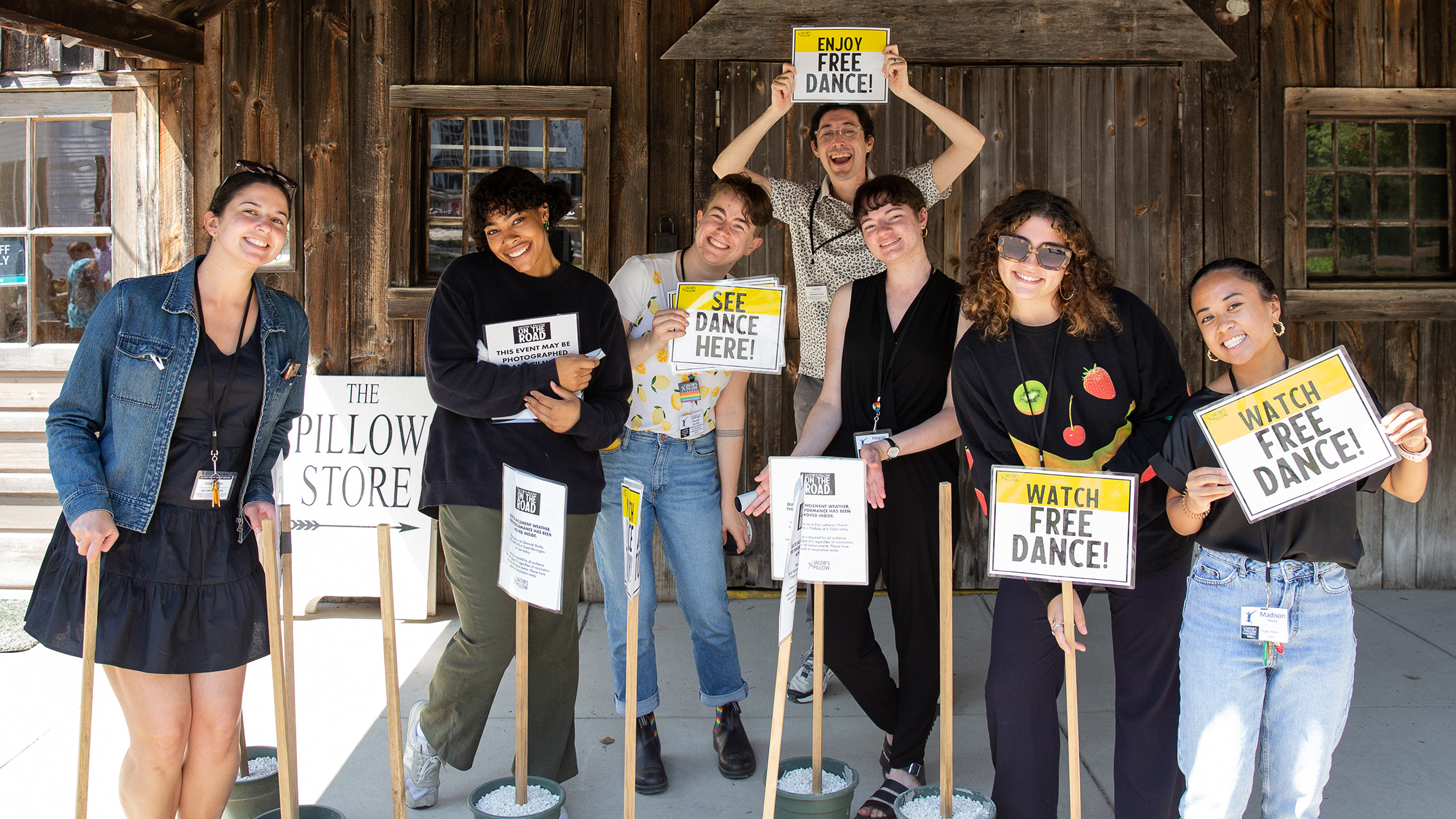 A group of people hold signs that say "Watch FREE Dance", "Enjoy FREE Dance", and "See Dance Here" designed in black and yellow. They are all standing in front of a wood building.