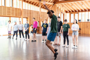 Sterling Harris, wearing tap shoes, balances on his toes and raises his arms above his head. 10 dancers look on.