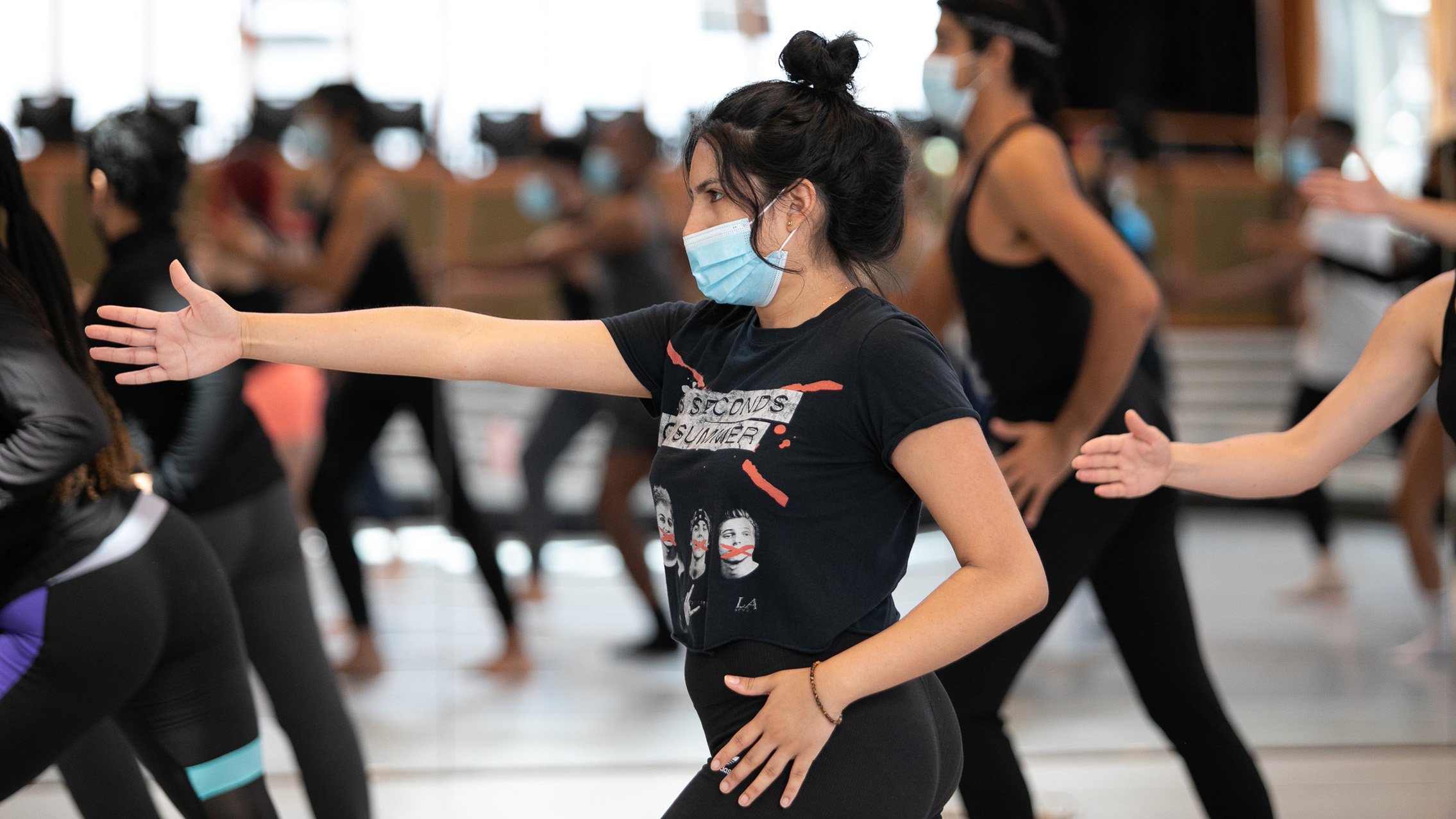 A dancer in a class reaches her right arm forward with her left hand placed near her hip, left elbow pointing backward.