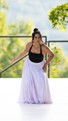A dancer on an outdoor stage sings into a head microphone. The dancer leans forward with one hand on a hip gathering a long white skirt and the other reaching backward.