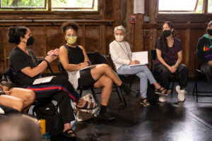 Choreography Fellows, Risa Steinberg, and choreography Yin Yue sit in a circle in folding chairs, wearing masks and talking to one another.