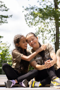 A young girl with dark hair smiles and puts her arms around a woman with a dark ponytail. Both are seated on an outdoor stage and wearing brown t-shirts.