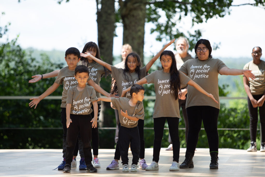 A group of children wearing brown Dance for Social Justice t-shirts stand on an outdoor stage with their arms outstretched.