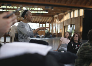 Dianne McIntyre sits in the Perles Family Studio and addresses the Choreography Fellows sitting on the floor in front of her. She gestures with her hands while speaking to them.