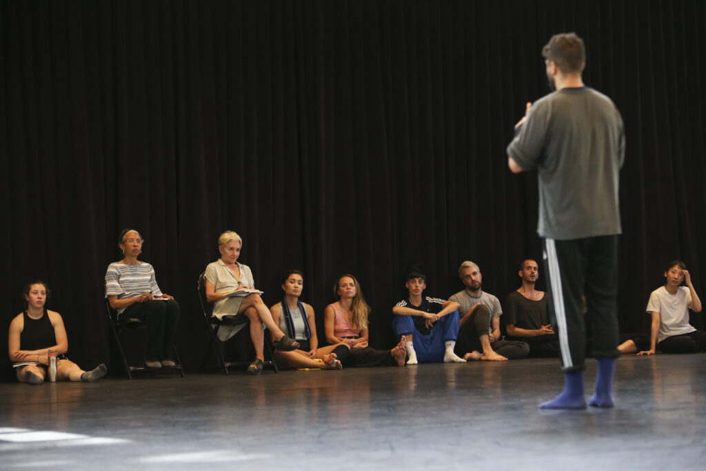 A dancer faces Dianne McIntyre and Risa Steinberg, their back to the camera. Other dancers and choreographers sit on the floor on either side of Risa and Dianne.