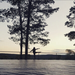 The silhouette of a woman stands on the outdoor Henry J. Leir Stage at dusk. She is extending her right leg to the side and reaching her right arm outwards.