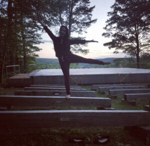 A woman stands on a bench in front of an outdoor stage at dusk. She is extending one leg behind her with the opposite arm reaching towards the sky.