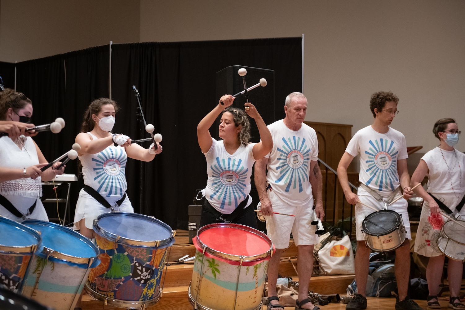 A group of six people stand, each playing their own drum. The woman in the middle holds two drumsticks above her head.