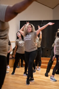 A woman with curly blonde hair dances with her arms above her head. She is wearing black pants and a shirt that says "Dance for Social Justice".