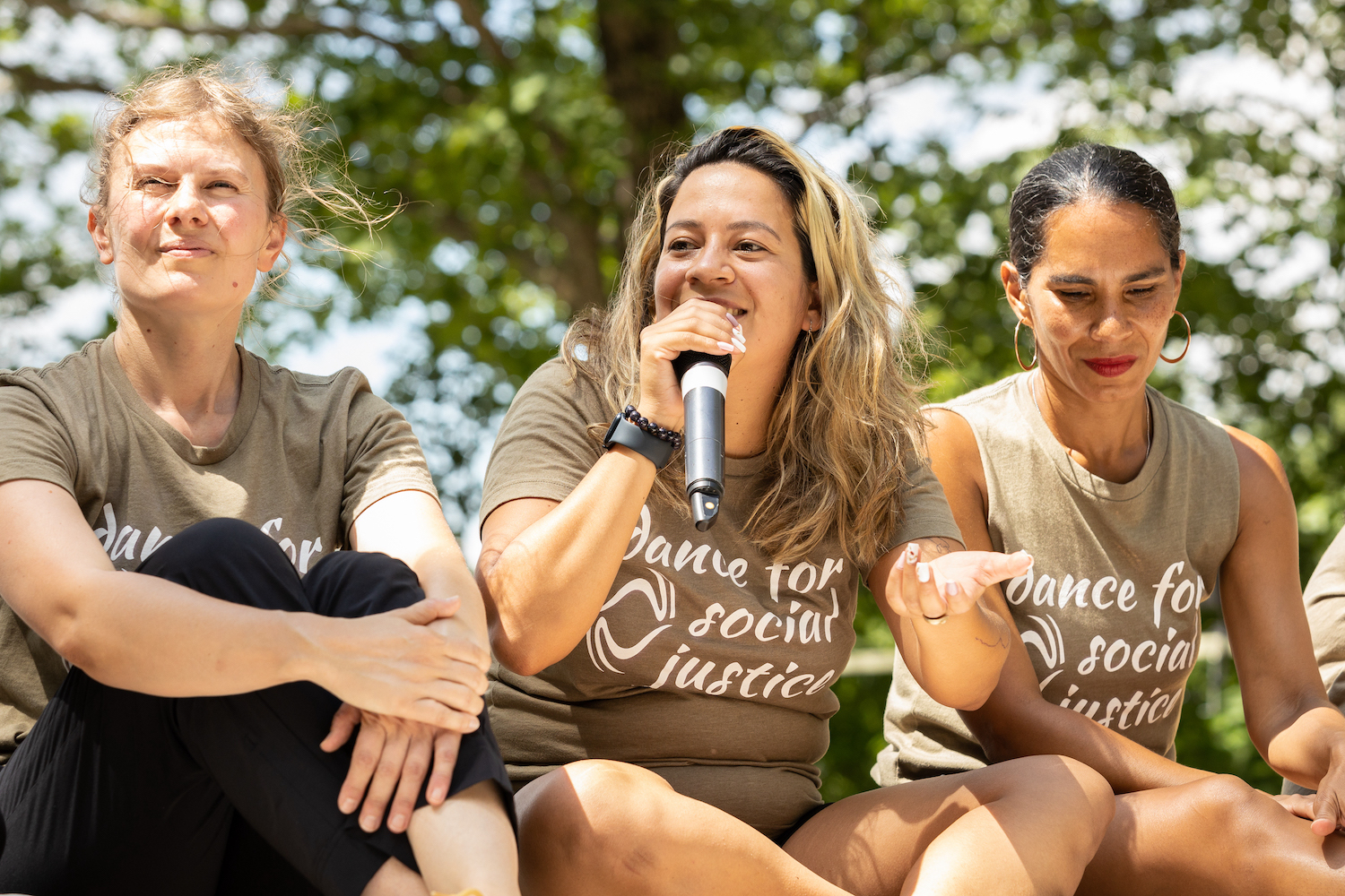 3 women sit cross legged with trees behind them. The one in the middle holds a microphone while she speaks.