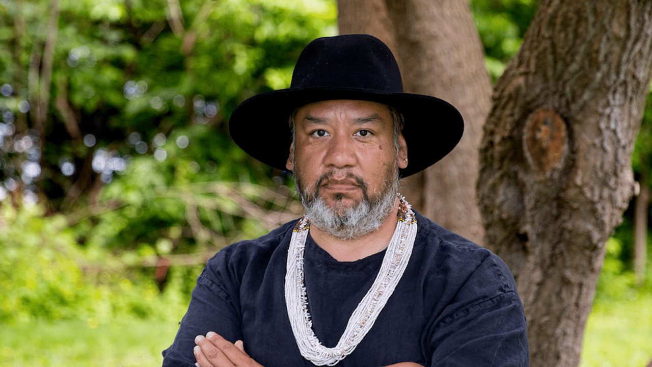 Jeffrey Gibson stands in front of a tree, wearing a black hat and a dark t-shirt while crossing his arms.