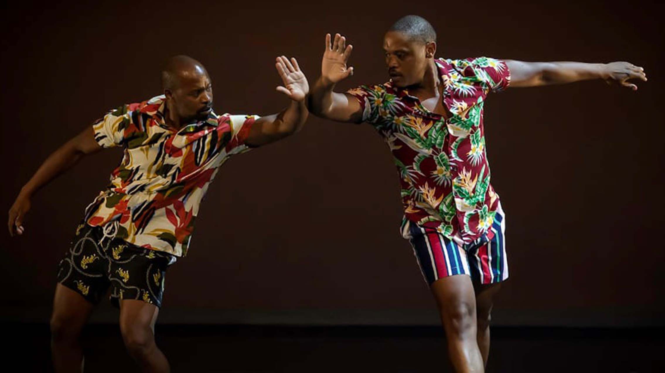 Two dancers in colorful outfits connect with their arms in a movement that is similar to a high five.
