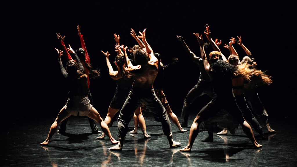 Martha Graham Dance Company dancers faced towards the back of the stage with their arms extended to a light coming from the back.