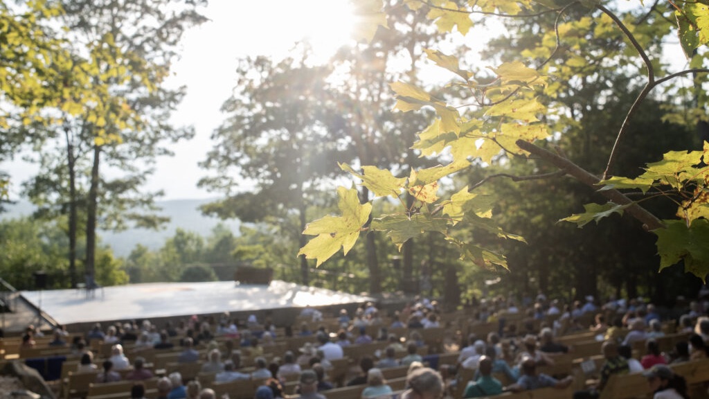 The sun shines through the leaves over the outdoor Henry J. Leir stage as a full audience looks on.
