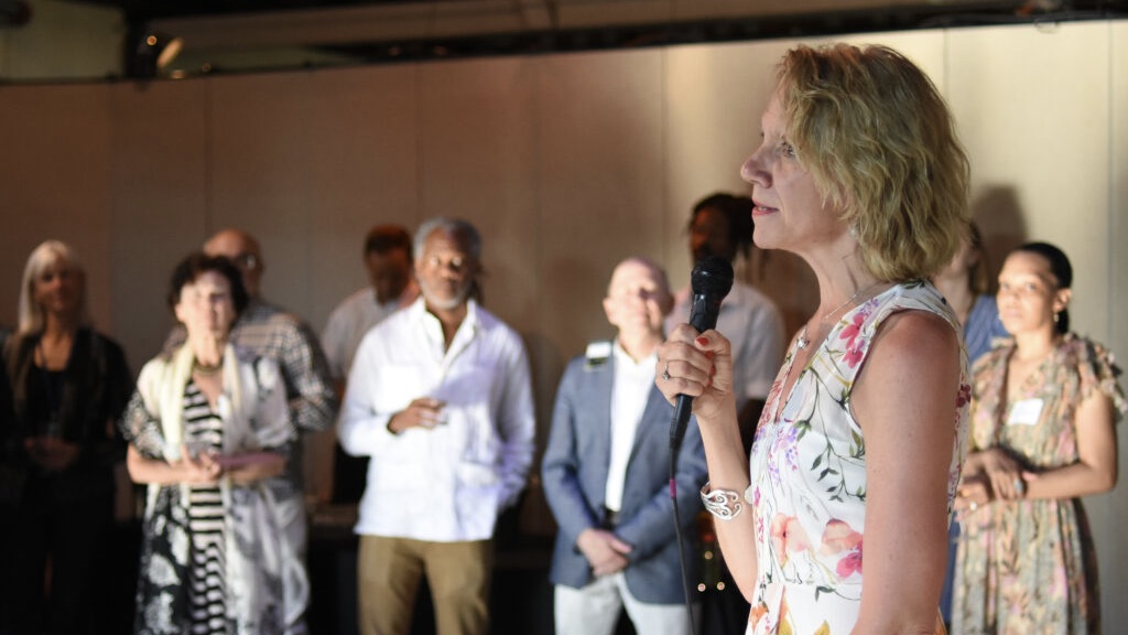 Pamela Tatge, a tall blonde woman, holds a microphone as group of people look on.