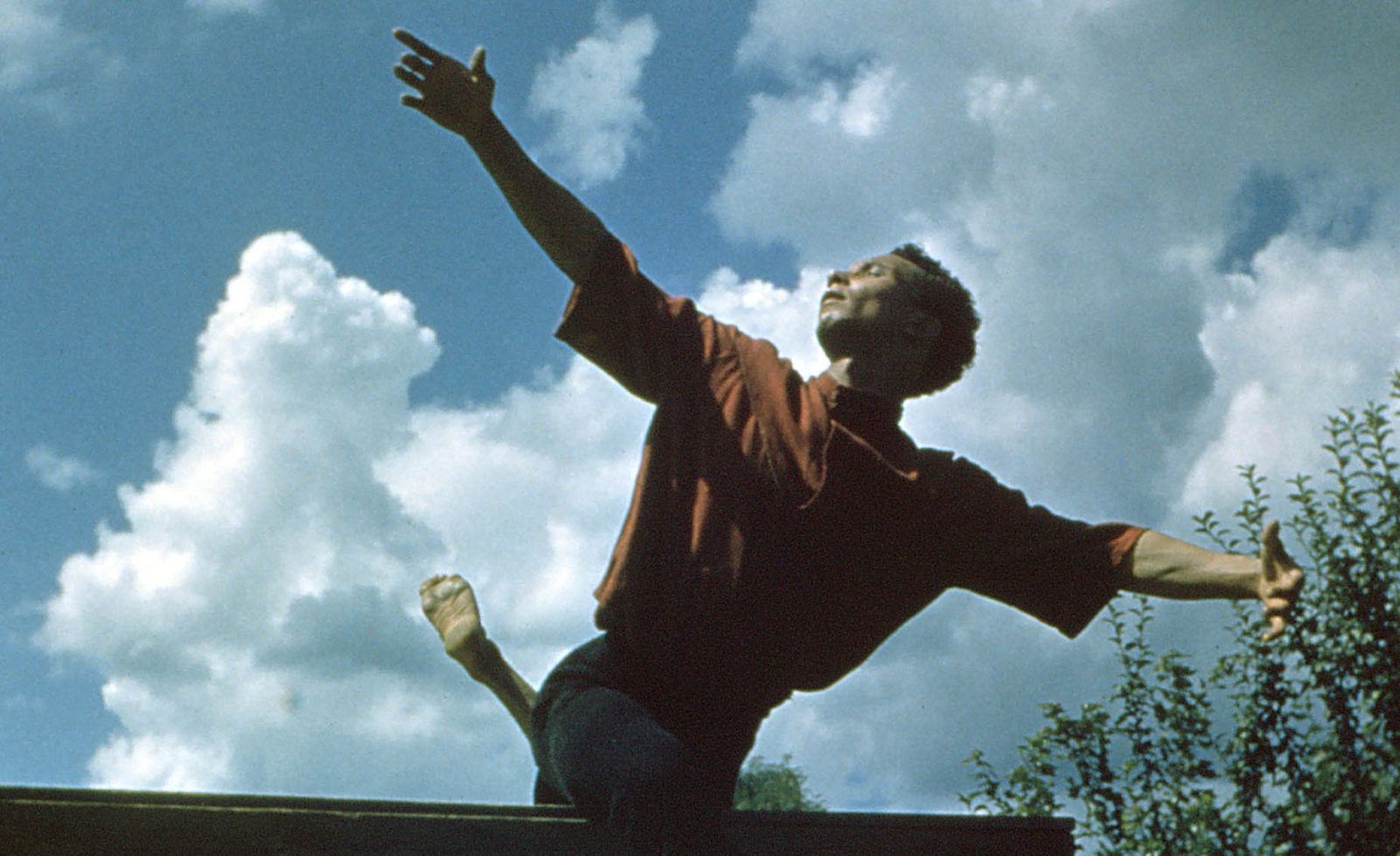 Talley Beatty, a black man, looks towards the sky and reaches one arm upward while sitting on a bench.