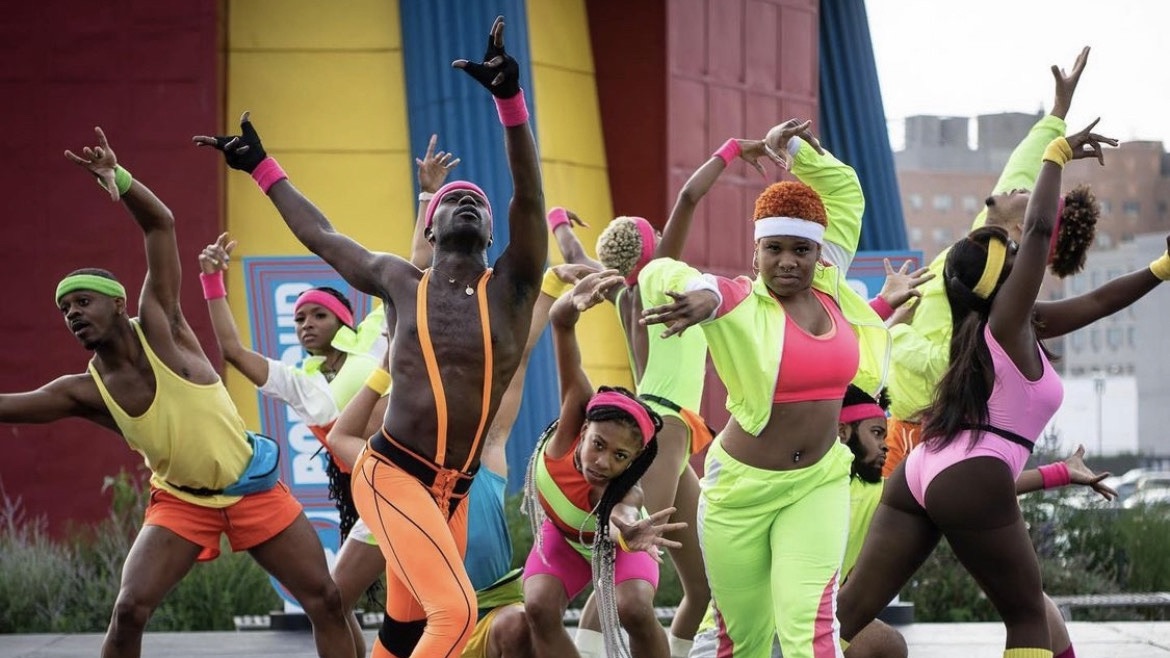 Multiple dancers outdoors wearing bright neon clothing reaching in multiple different directions.