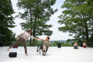 Image description: A diverse group of disabled and non-disabled dancers perform on a white outdoor stage, which opens up to a majestic background of trees, rolling hills and sky. In the foreground, Zara and Julie hold a counterbalance as Julie tilts to the side in her wheelchair, holding Zara’s arm as Zara balances with one lower limb extended. In the background, Alaja and Louisa kneel on the ground with one palm reaching outwards, among suitcase set pieces. The dancers wear period costumes in beige, green and maroon, with red and white flowers in their hair. 