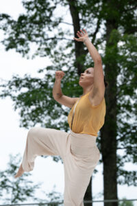 An Axis Dance member stands on one foot and holds both arms above her head. She looks upward to the sky.