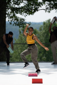 A dancer has one foot off the ground, a hand to her side, and the other hand above her head. She smiles towards the camera.
