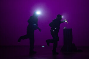 two dancers dance in almost blackout but purple haze. the two people are in all black with headlamps