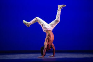A man does a handstand. He wears white pants and the background is illuminated blue.