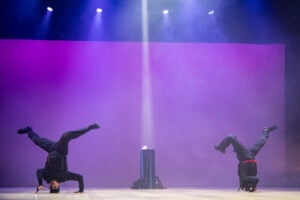 Two dancers do headstands next to each other. They both wear all black. There is a purple background behind them as well as a podium in-between them that is illuminated.