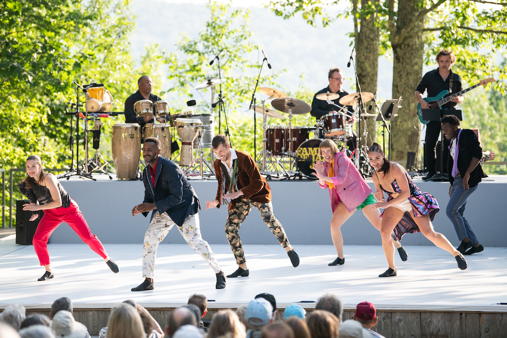 Photo of dancers leaning right and clapping. All of the dancers are smiling and wearing brightly colored clothing.