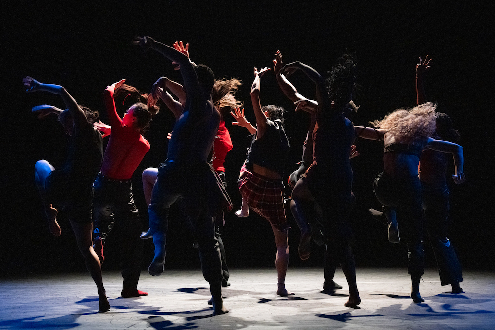 A photo of a group of dancers from behind. They all hold their arms up in the air and all stand on one leg.