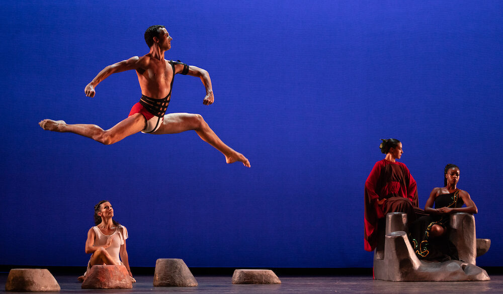 A photo of four people. Three of them sit, a man jumps. The man jumping looks proudly outward. A woman sitting down watches him, the other two women look away.