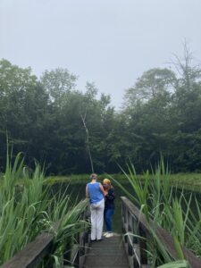 two people stand by a pond. one is wearing a blue shirt and the other a bright yellow bandana. The boardwalk in the foreground is framed in reeds, and the air is misty