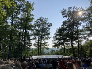 A photo of the Henry J. Leir stage. Tall trees rise above a gray Marley stage, audience members sit in the foreground. The sun is high in the sky and there isn't a single cloud in site