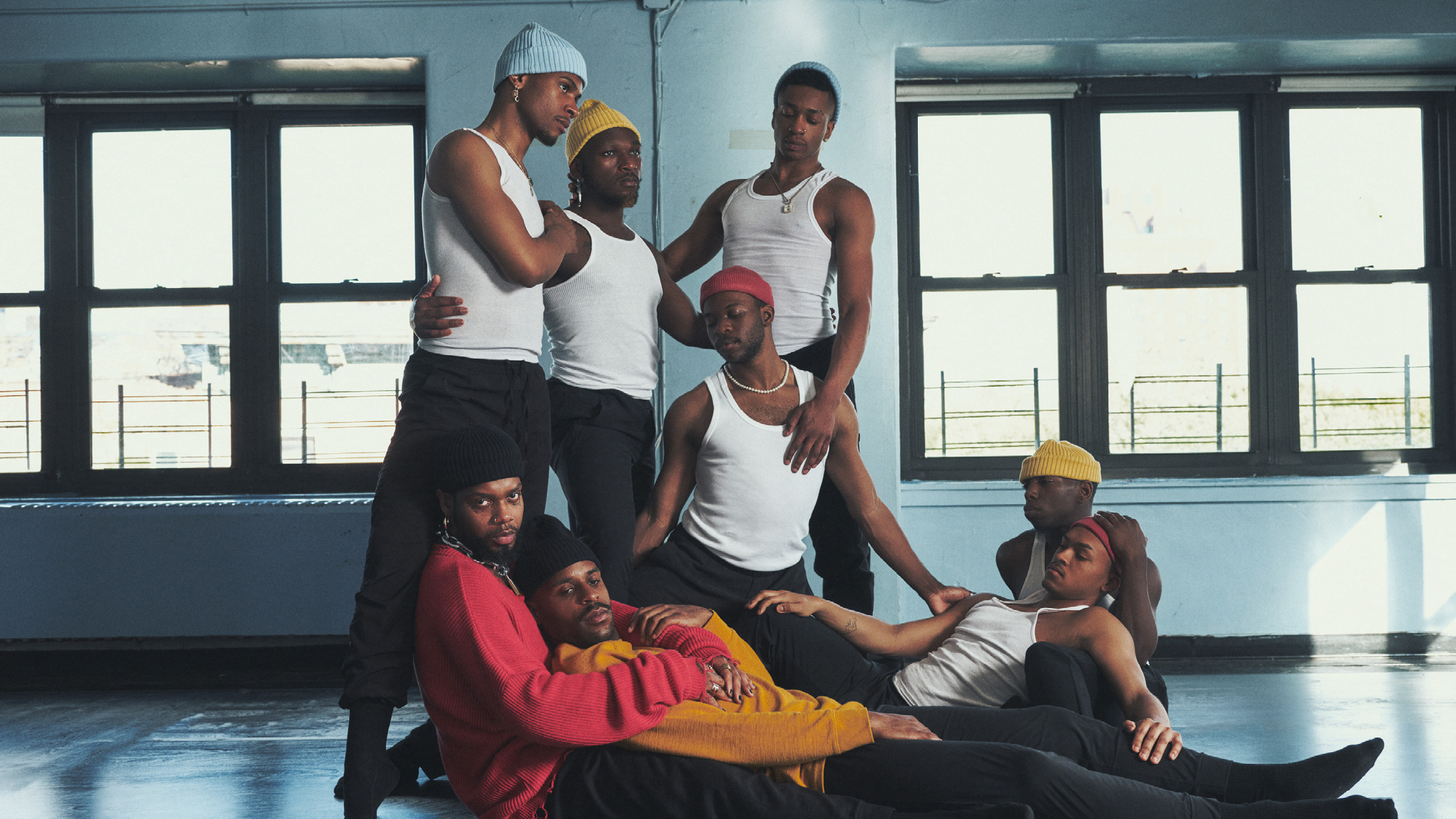 A group of black men sit and stand in close proximity to one another, all touching the shoulder or back of the men by their sides. All of them are wearing beanie hats, black pants, and white tank tops or colorful crewnecks.