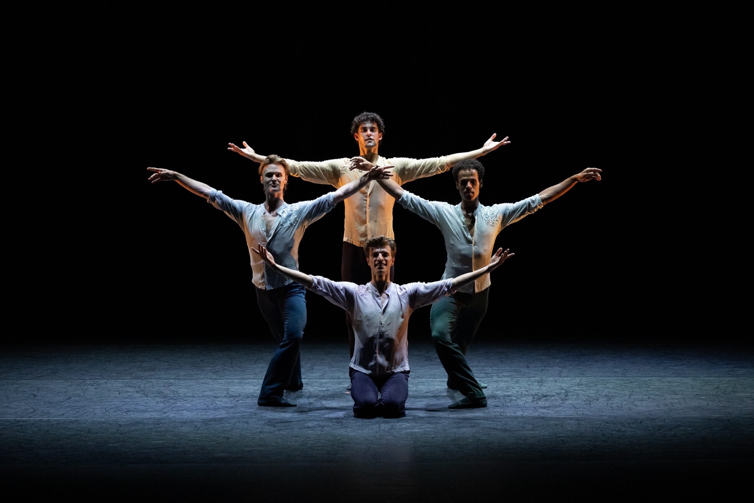 Four men dancers pose together in a dim spotlight that casts soft shadows across their faces and costumes. The rest of the stage is dark. They wear matching costumes of long pants and button down shirts, but each wears a different muted color. One dancers kneels on front, two lunge on either side of him, and a final dancers stands in back, all with their arms extended out to the side. It creates a symmetrical formation.