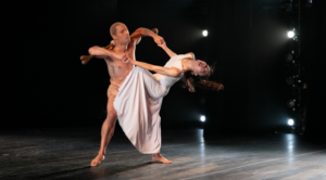 A man in nude colored attire and a woman in a long white dress dance together dramatically on a dark stage.