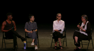 Four people sit in chairs on stage, and the three people sitting on the left look to the right as the woman on the far right is talking into a microphone.