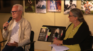 A man and a woman sit in chairs in front of a photo galler, talking into their microphones.